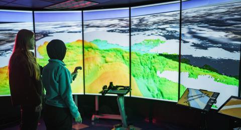 两名研究人员站在一个墙壁大小的屏幕前，屏幕上显示着海洋测绘数据